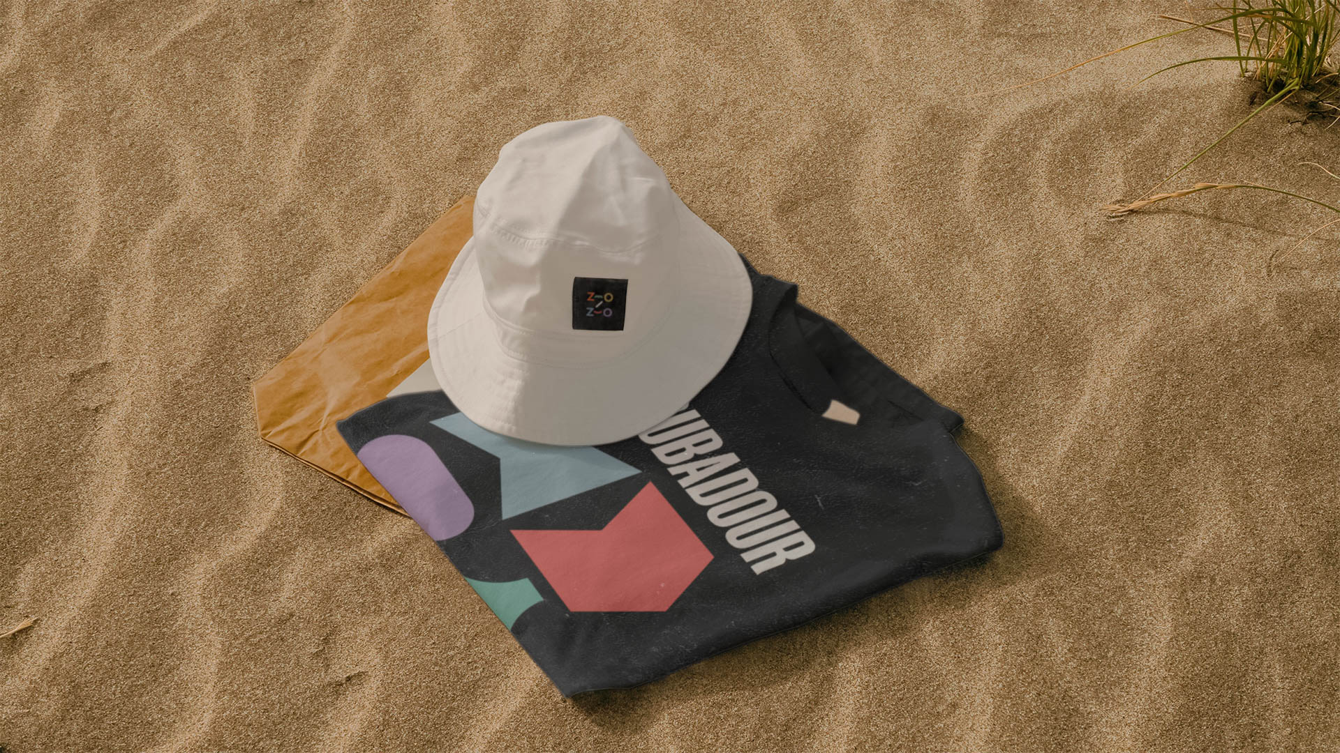 A white bucket hat with a black square patch on the front rests on a dark-colored t-shirt with a geometric pattern in teal, purple, red, and green. The t-shirt has the word “Jubadour” printed in white across the chest. The hat and shirt are placed on a light brown sandy surface.