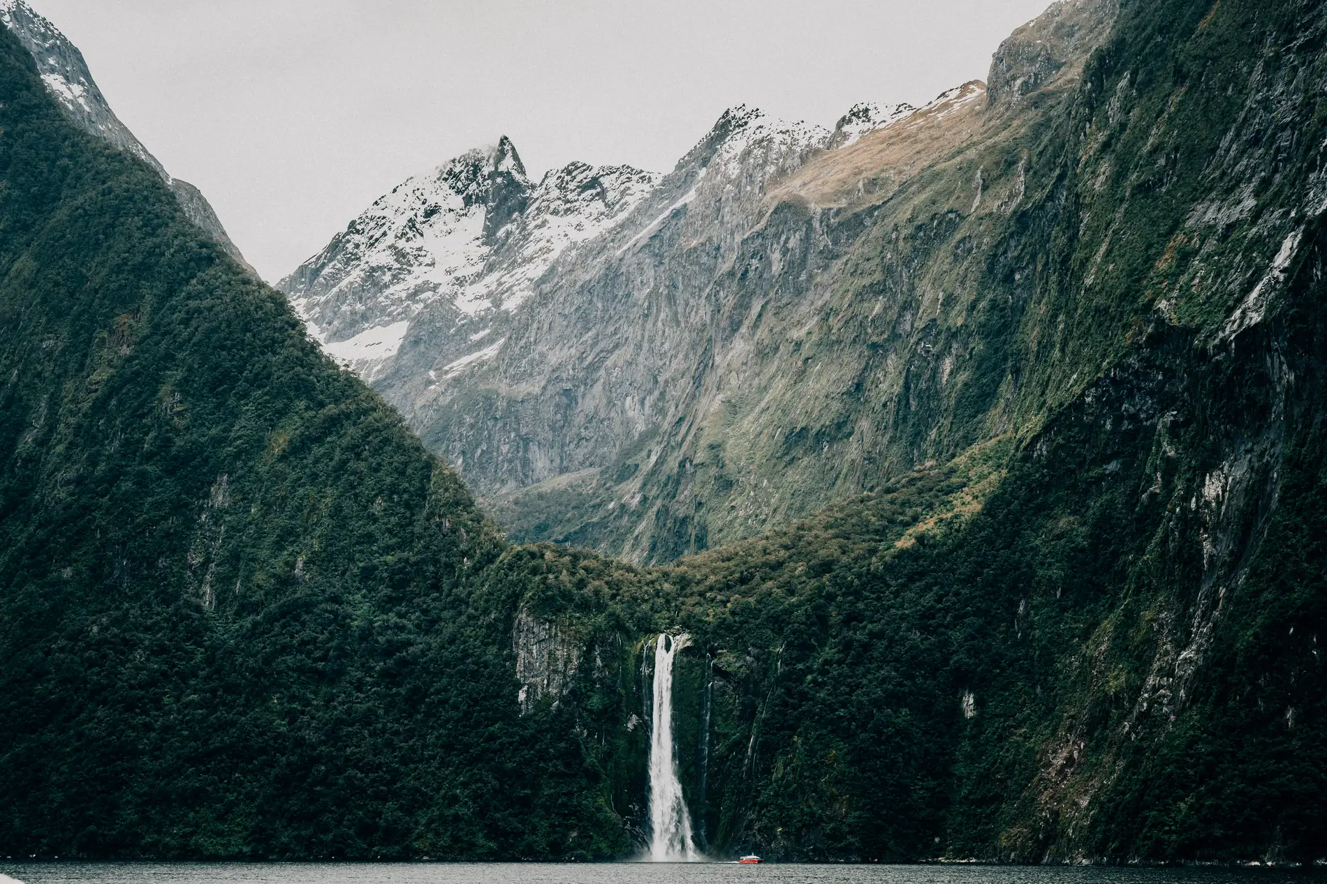 A tall waterfall dropping down a sheer, forested fiord wall below snow-covered peaks