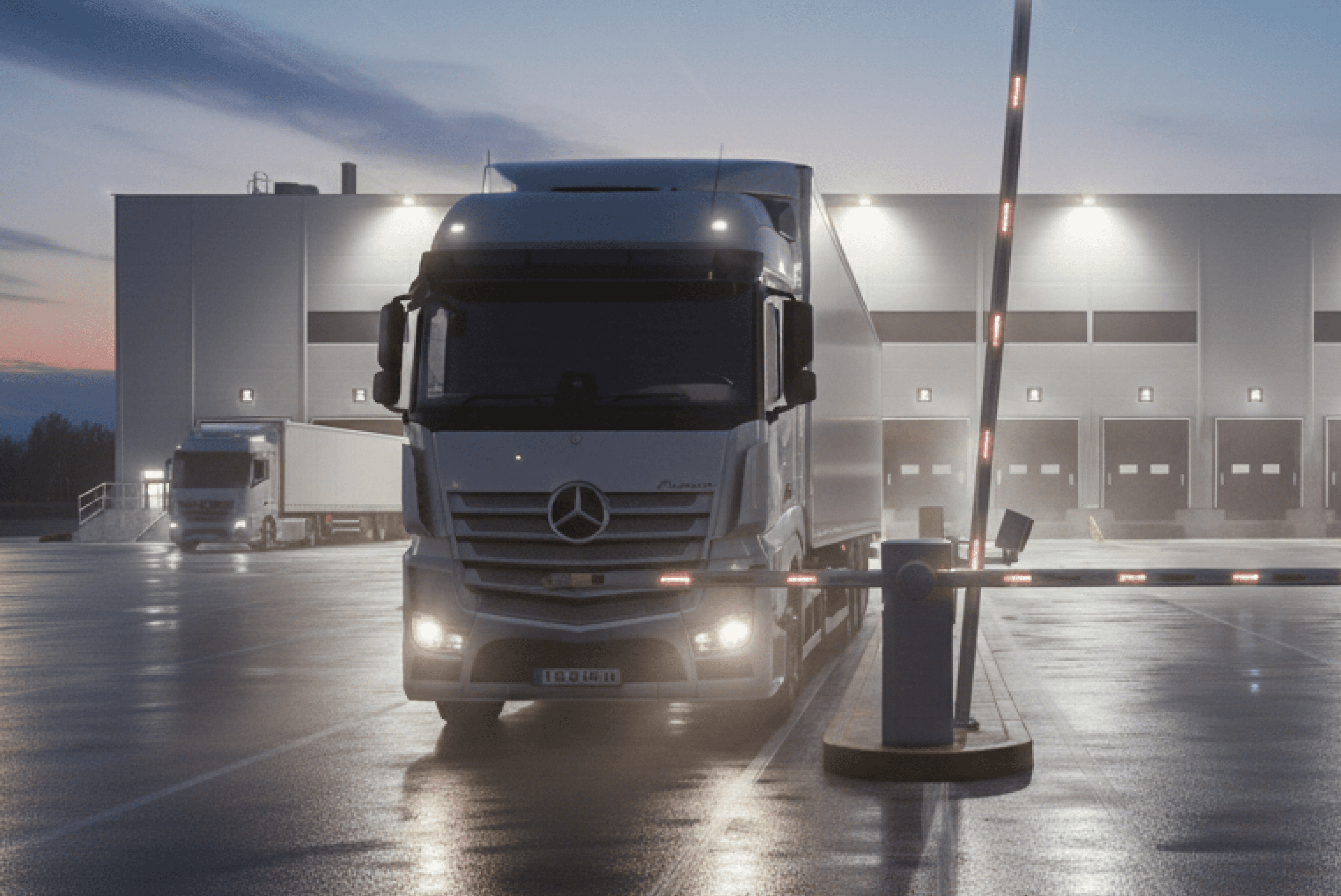 새벽 물류센터 야외 주차장에서 대형 화물 트럭(메르세데스-벤츠 악트로스)이 차단기 앞에 정차해 있으며, 배경에 대형 창고 건물과 또 다른 트럭이 보이는 이미지입니다.