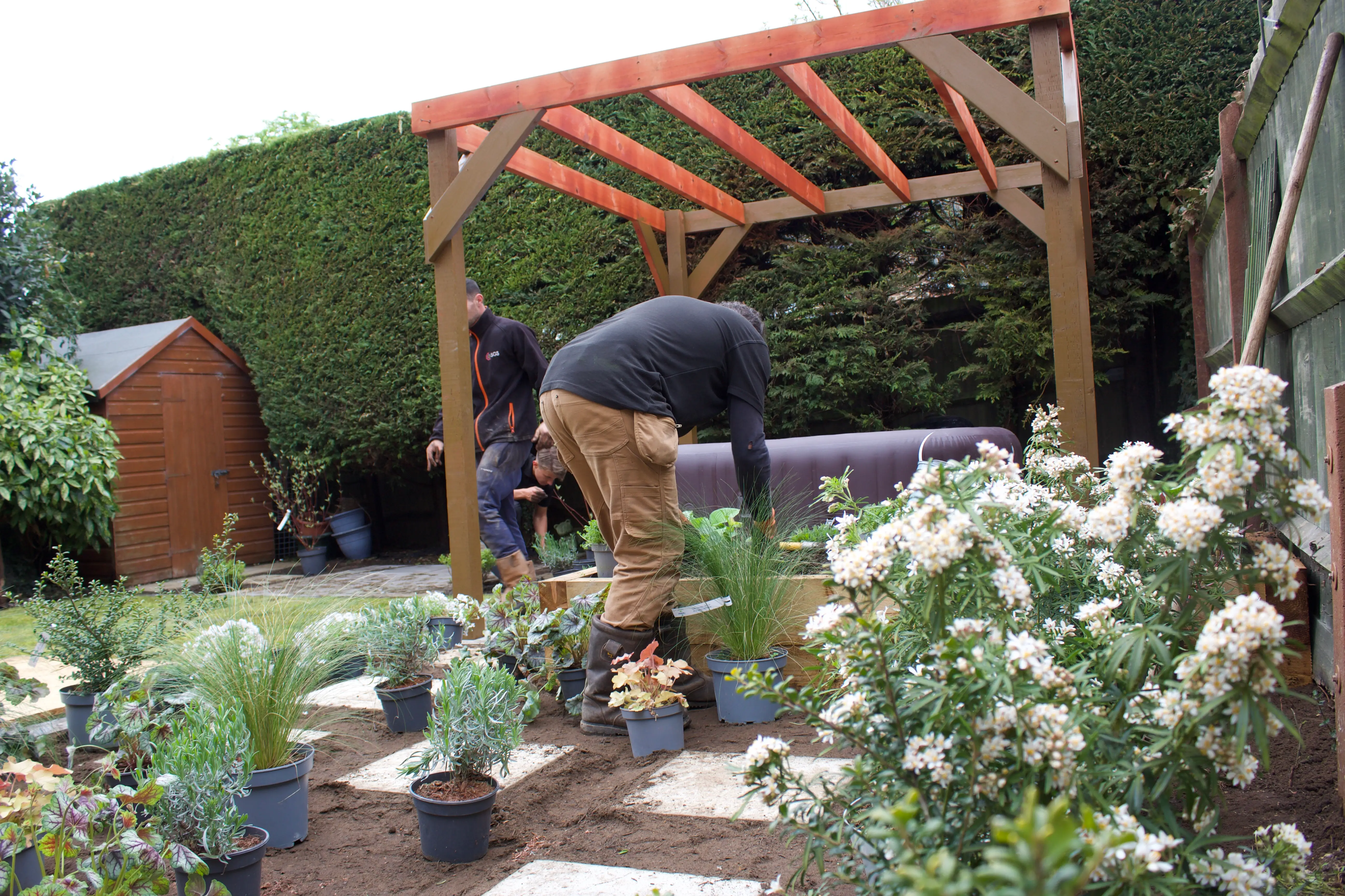 Person tending to plants under a wooden gazebo in a garden filled with various potted plants.