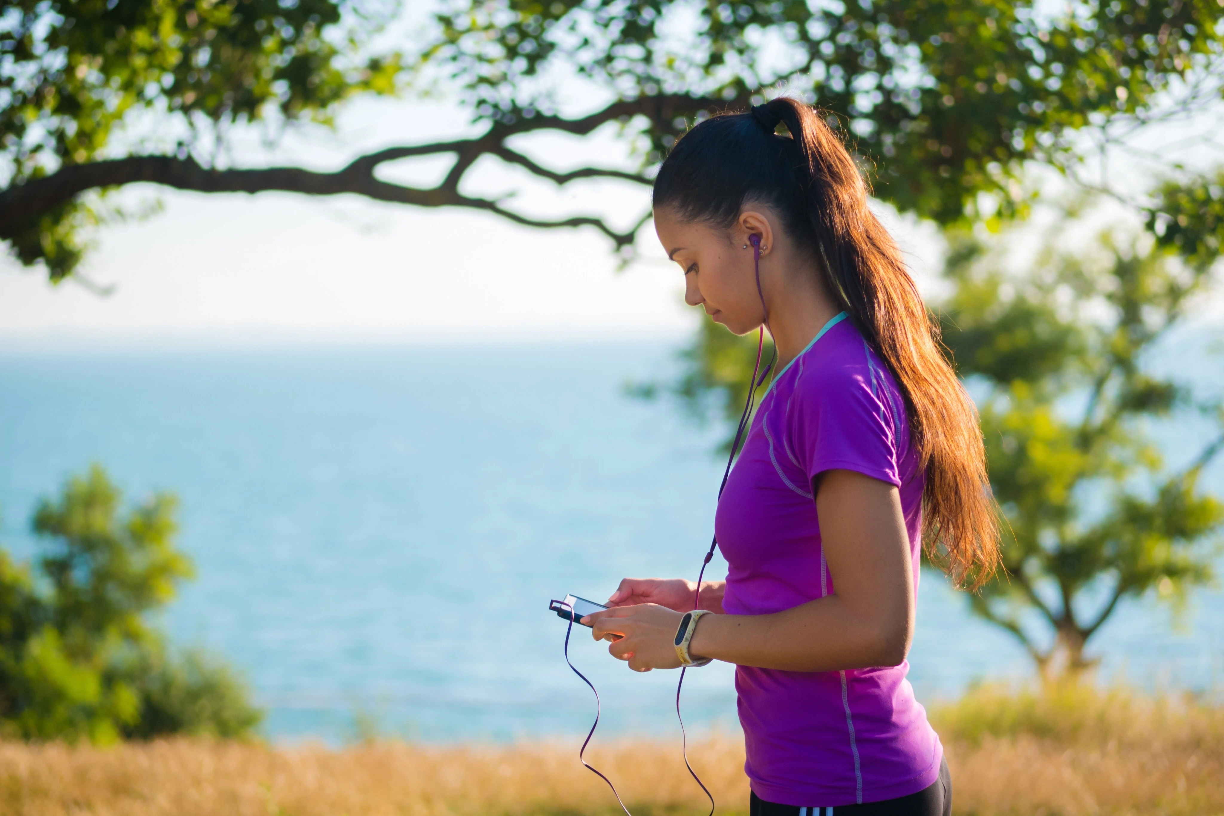 Mujer con ropa deportiva mirando su celular y auriculares puestos mientras camina junto al mar en un entorno natural.