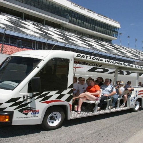 En gruppe mennesker kører i en sporvogn mærket "Daytona International Speedway" med tribunen i baggrunden.