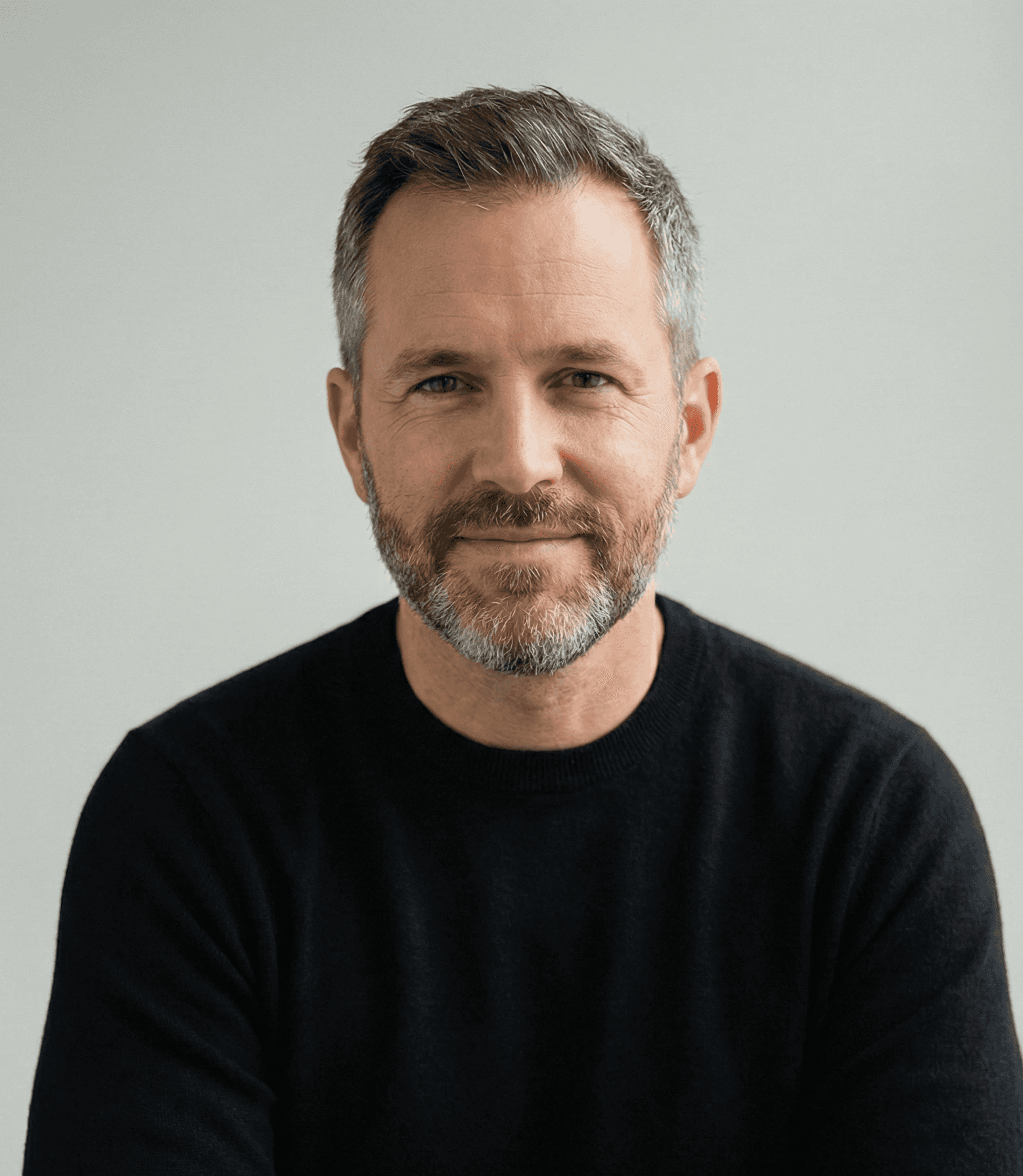Mature man with glasses and a trimmed beard smiling confidently, standing in warm natural light against a modern backdrop.