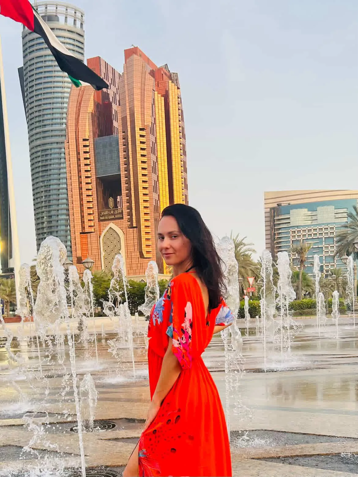 Woman in a red dress standing near a fountain, with modern high-rise buildings in the background, looking over her shoulder toward the camera.