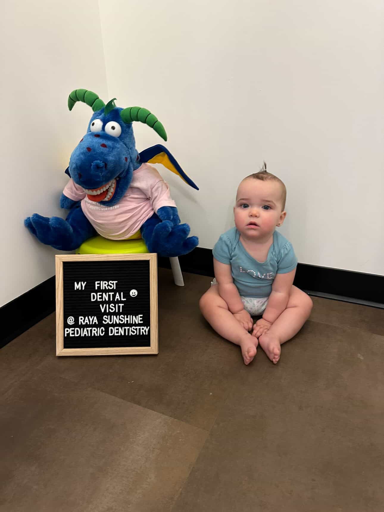 A baby sitting on the floor next to a blue dinosaur mascot holding a “My first dental visit” sign