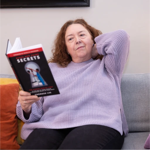 Older woman relaxing on a couch while reading a book titled 'Aches and Pains Secrets', rubbing her neck in pain