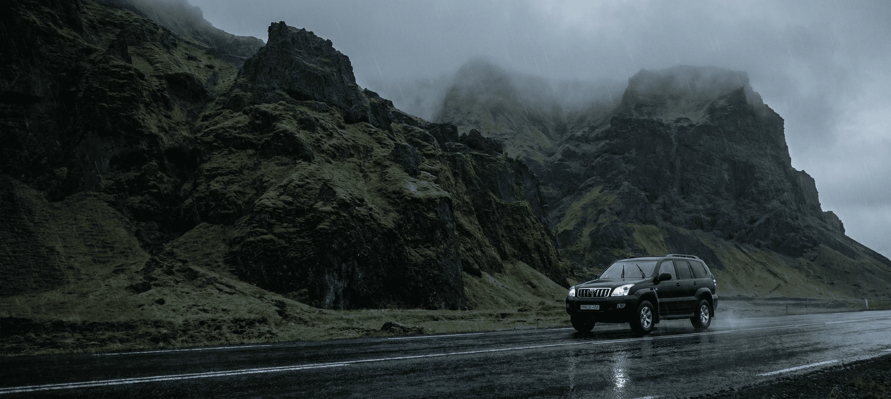 A dark SUV driving along a wet coastal road next to steep, moss-covered green cliffs.