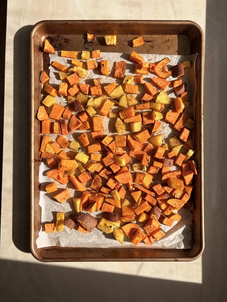 Roasted cubes of butternut squash, acorn squash, and sweet potatoes spread across a sheet pan, caramelized and lightly browned, coated in olive oil with sage and nutmeg.
