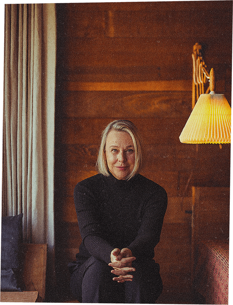 Headshot of a woman with straight blonde shoulder length hair smiles slightly, sitting in a wood paneled room, leaning forward with her hands clasped.