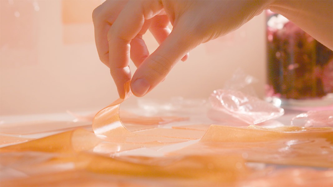 A hand is holding a piece of translucent, orange-hued material between its thumb and index finger. The material is thin and curved, partially folded and spread out on a surface. In the background, a blurred object with pink and purple tones is visible, along with a clear glass jar containing dark liquid. The scene is illuminated with warm, soft lighting.