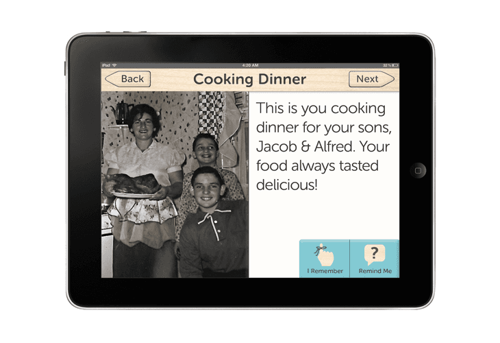 An iPad displays a webpage titled “Cooking Dinner” with a photo of a family in a kitchen setting. The text reads, “This is you cooking dinner for your sons, Jacob & Alfred. Your food always tasted delicious!” Below the text are two buttons: “I Remember” and “Remind Me.”