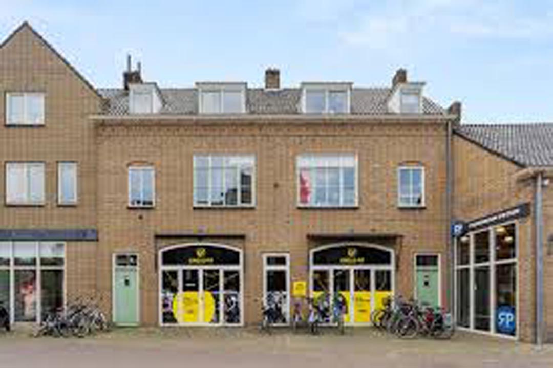 A two-story brick building with gray roofs, large windows, and yellow doors, under an overcast sky.