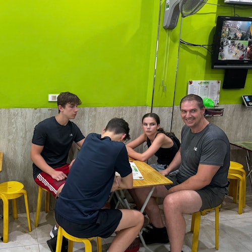 Fire personer sidder ved et lille bord med gule taburetter i et lyst oplyst rum med limegrønne vægge og et vægmonteret TV.