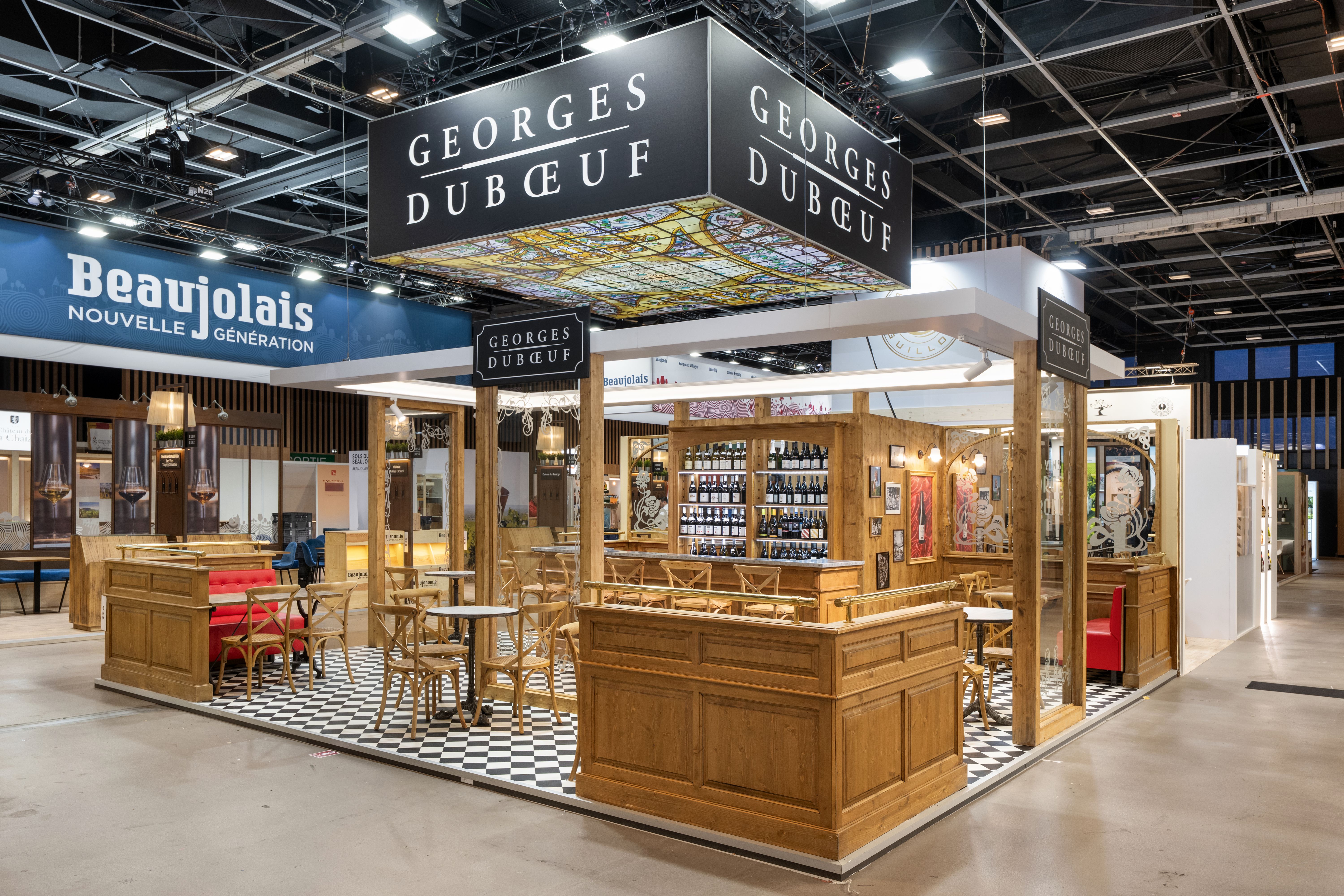 Stand Georges Dubœuf Wine Paris 2026 réalisé par Expace – vue frontale avec façade mur de bouteilles en bois massif, caisson logo doré suspendu et comptoir dégustation Beaujolais