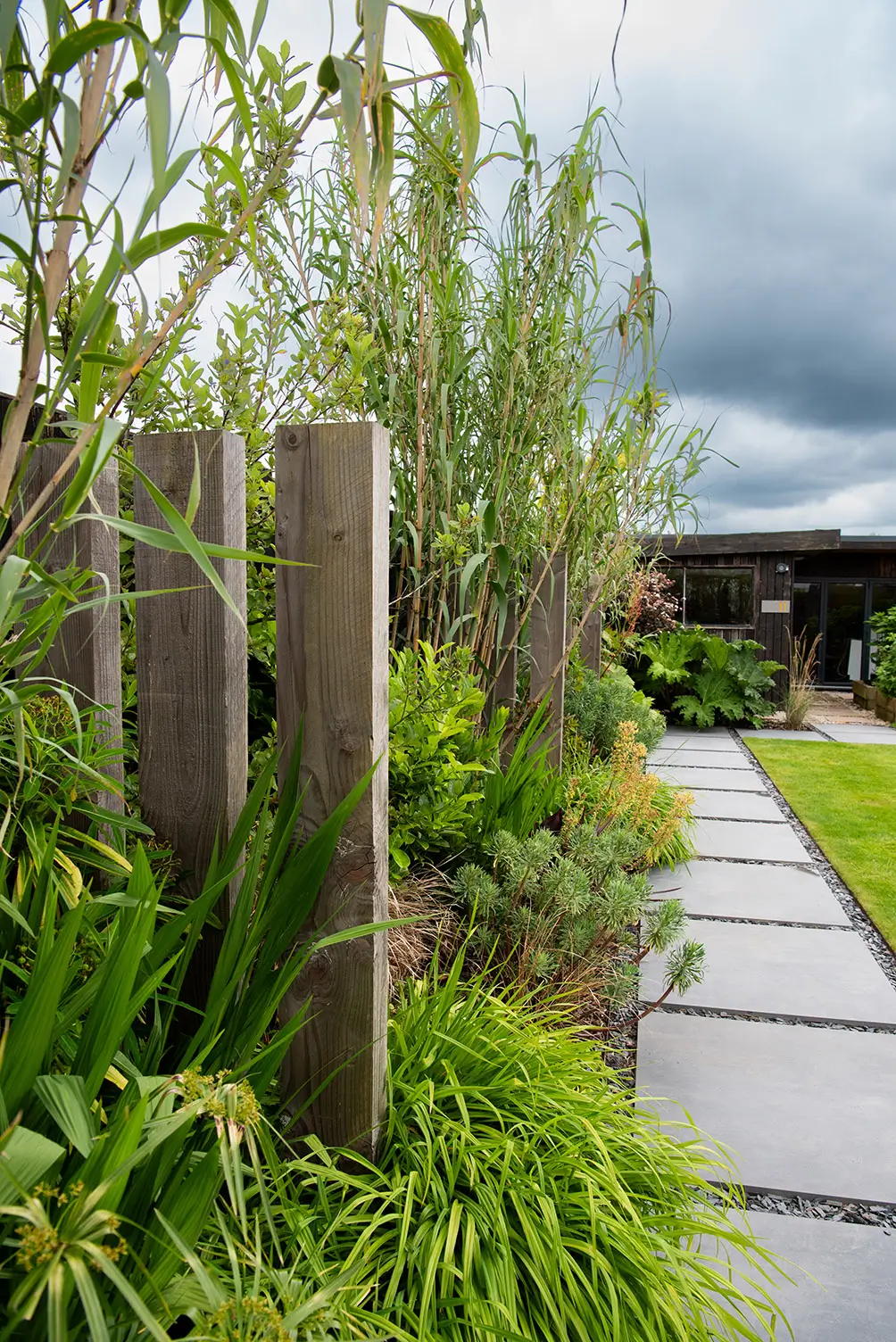 Lush green plants line a stone pathway, leading to a building under a cloudy sky.