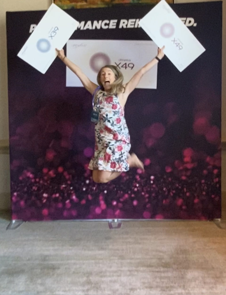 A person in a floral dress is enthusiastically jumping with arms raised, holding two white signs, against a vibrant backdrop with the text "PERFORMANCE REIMAGINED," conveying excitement and energy.