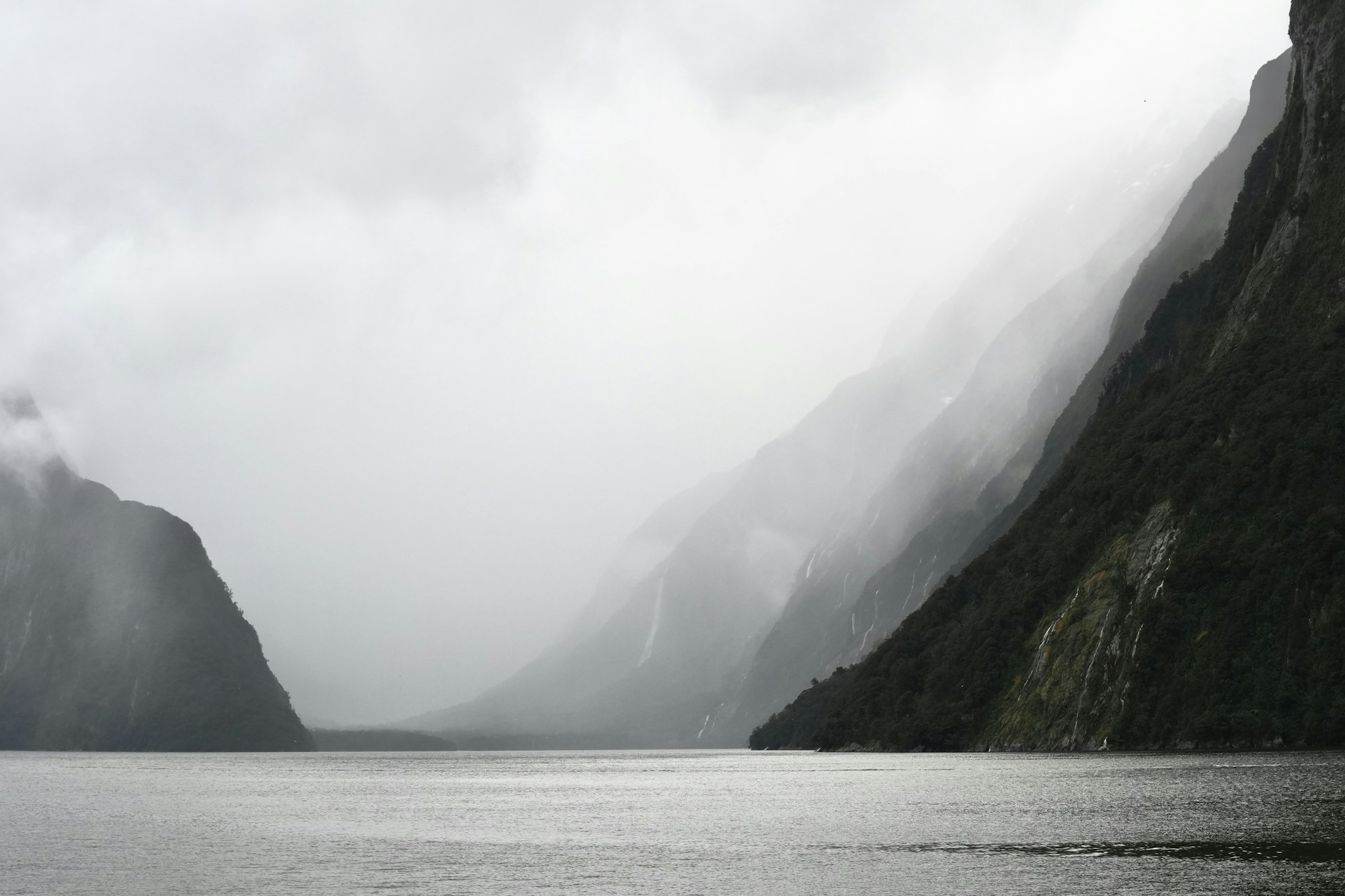 Steep mountains rising from dark water under a heavy mist and overcast sky