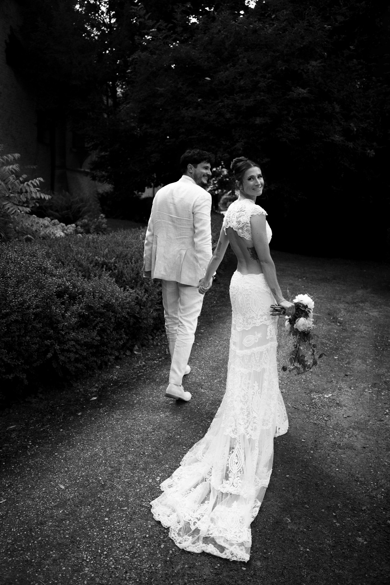 Sposi camminano su viale alberato della Rocca di Soragna in bianco e nero, lei con bouquet, reportage matrimoniale