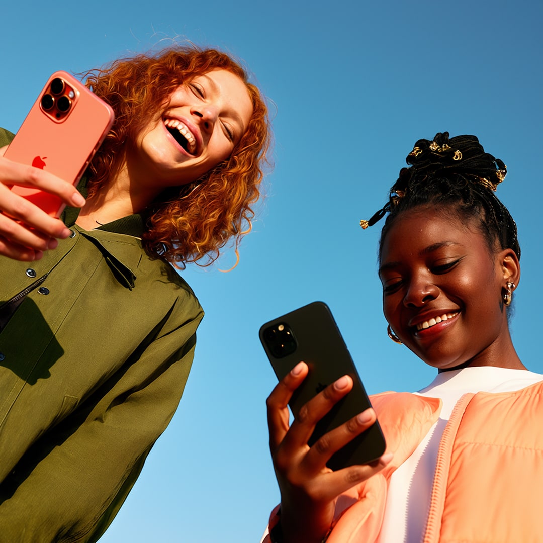 Two young women outdoors laughing and looking at their smartphones against a bright blue sky, used as hero photography for the NCBA brand.