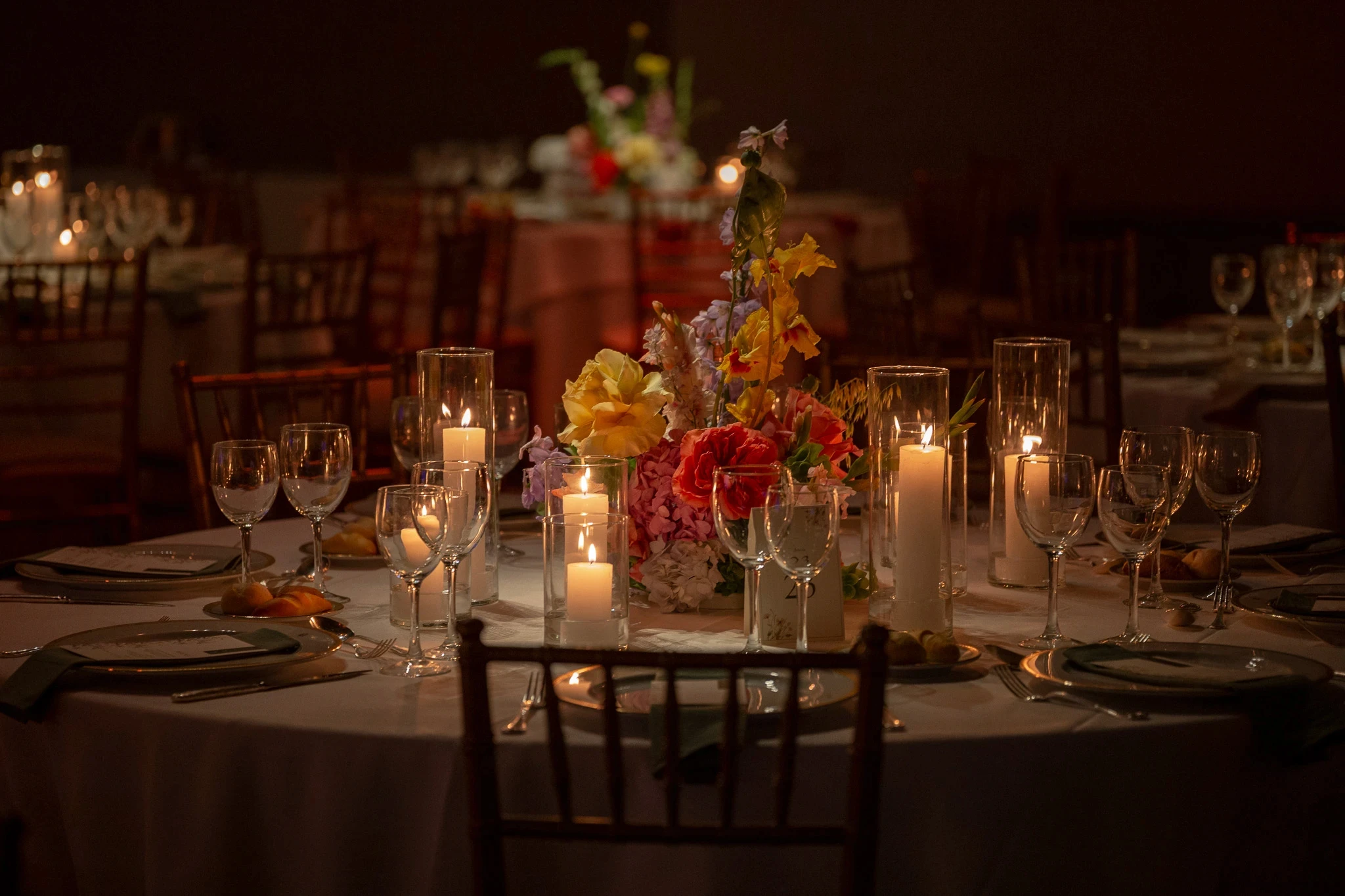 Mesa de recepción con centro floral coral y amarillo, rodeada de velas cilíndricas en fanales de vidrio.