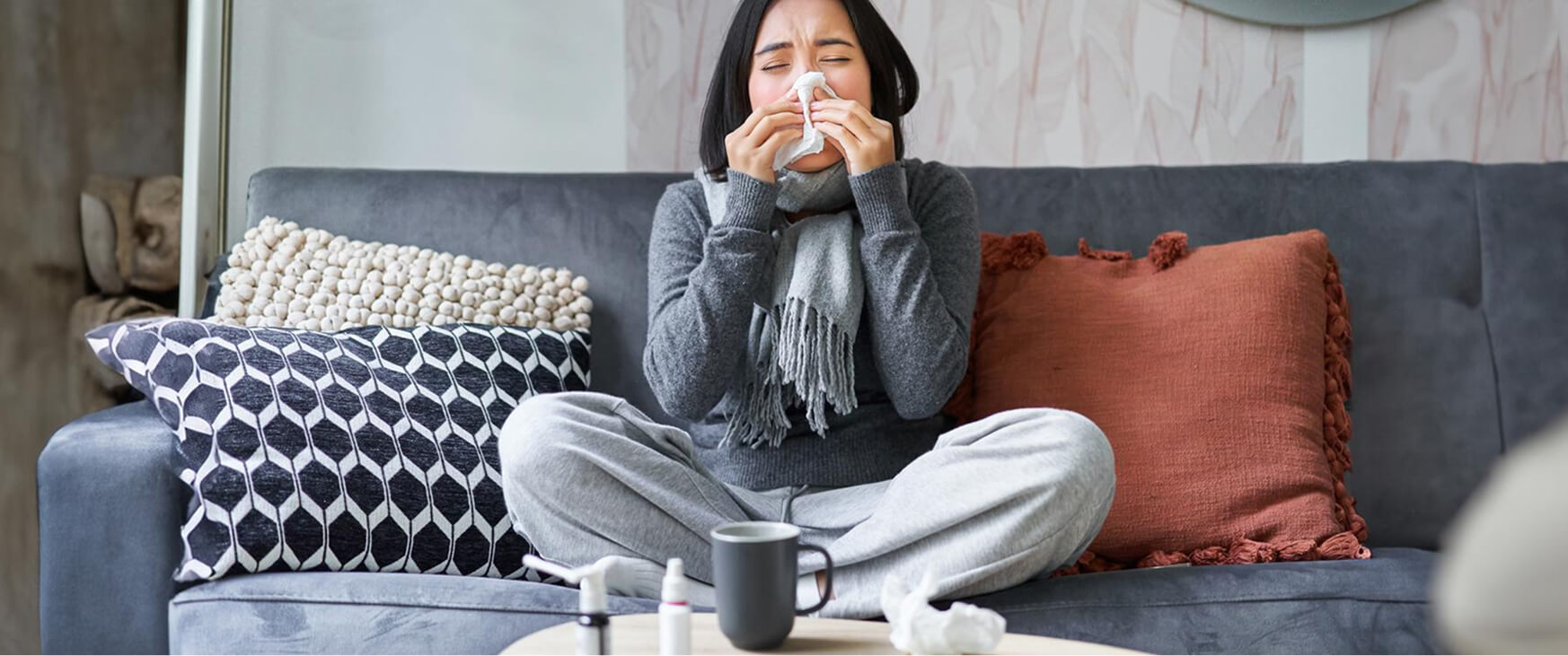 A woman sitting cross-legged on a gray couch sneezes into a tissue, surrounded by cozy pillows, with nasal spray and a mug on the coffee table, illustrating the need for ayurvedic remedies for allergies.