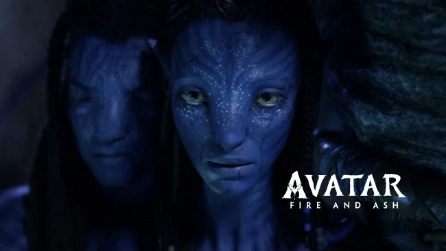 A blue-skinned alien with bioluminescent patterns stares intently, partially illuminated in a shadowy setting with another figure in the background, accompanied by the text "Avatar: Fire and Ash" on the lower right.