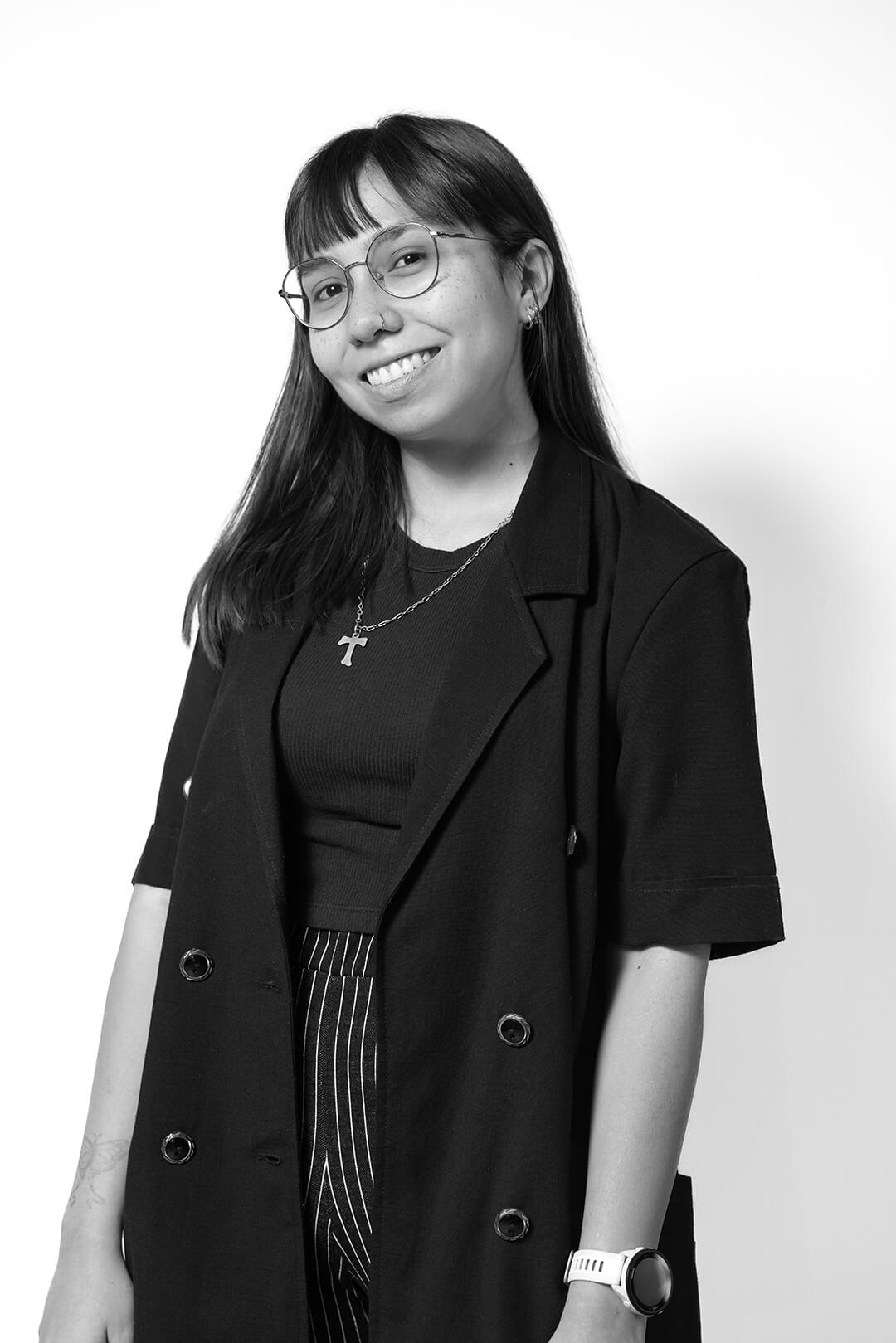 Black and white portrait of a person smiling, wearing glasses and a cross necklace. They have long hair and are dressed in a black jacket and striped pants.