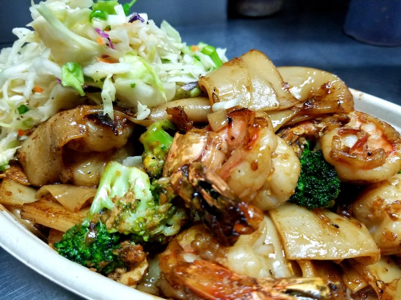 A close-up view of stir-fried wide chow fun noodles, wild shrimp, and broccoli coated in a dark sweet soy sauce, served alongside a portion of fresh cabbage slaw.