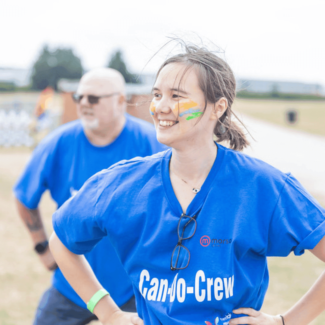 A smiling woman with face paint in green and yellow stands outdoors with her hands on her hips, wearing a blue "Can-Do-Crew" T-shirt. A man in a similar blue shirt is visible in the background.