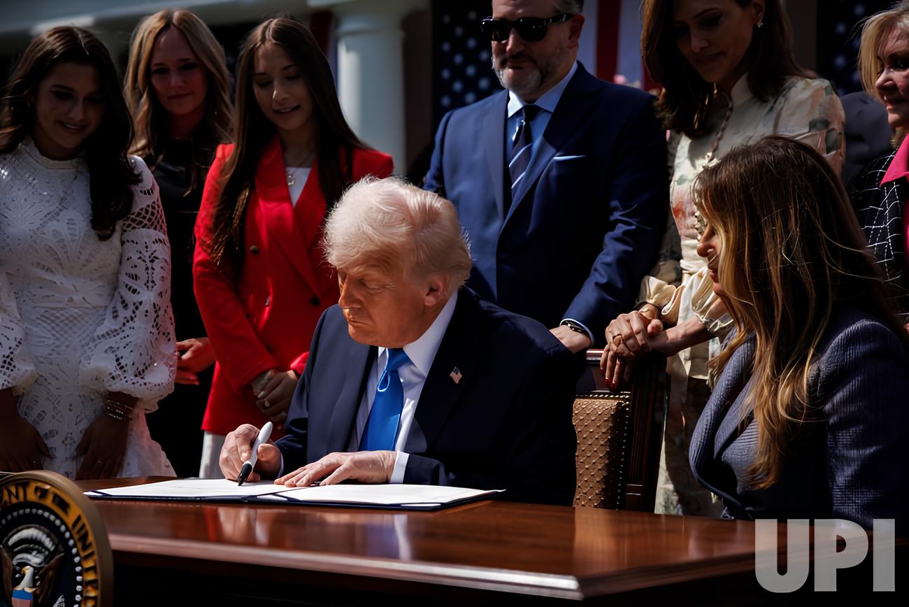 A formal signing ceremony: a suited official sits at a large desk and signs a document while a group of people stand around watching. The setting and staging suggest an executive or government action being finalized in public.