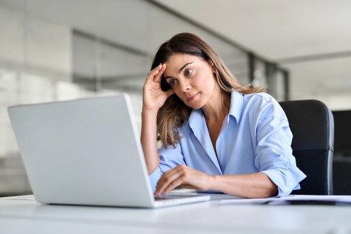 A woman in a light blue shirt sitting at a desk with a laptop, resting her head on her hand with a thoughtful or concerned expression