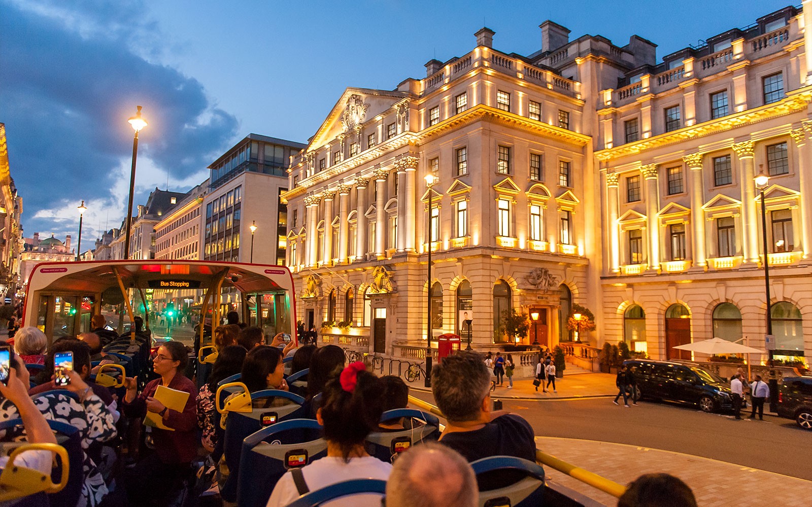 Open-top bus tour passing illuminated buildings in London at dusk.