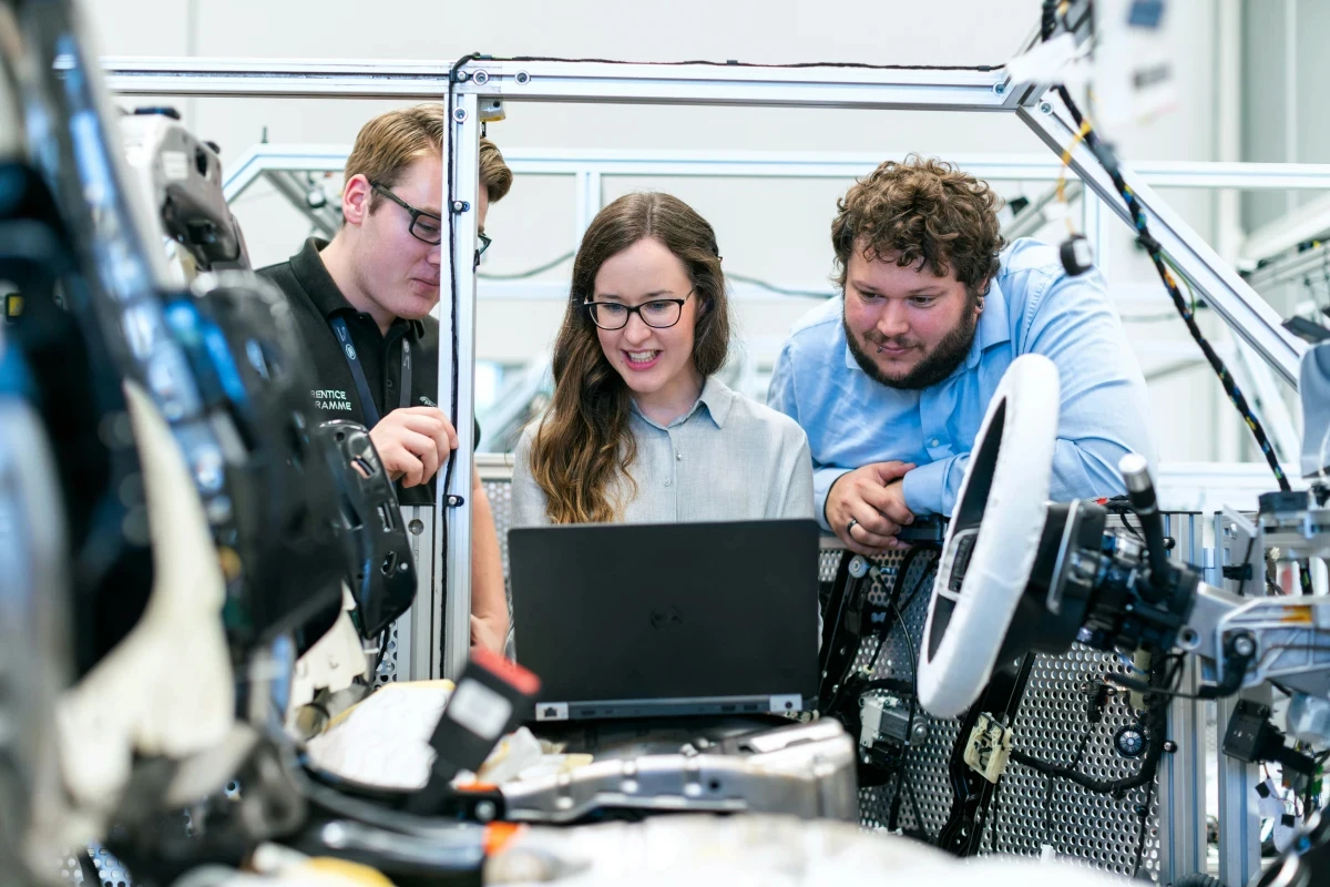 Three people look at a laptop beside a car part.