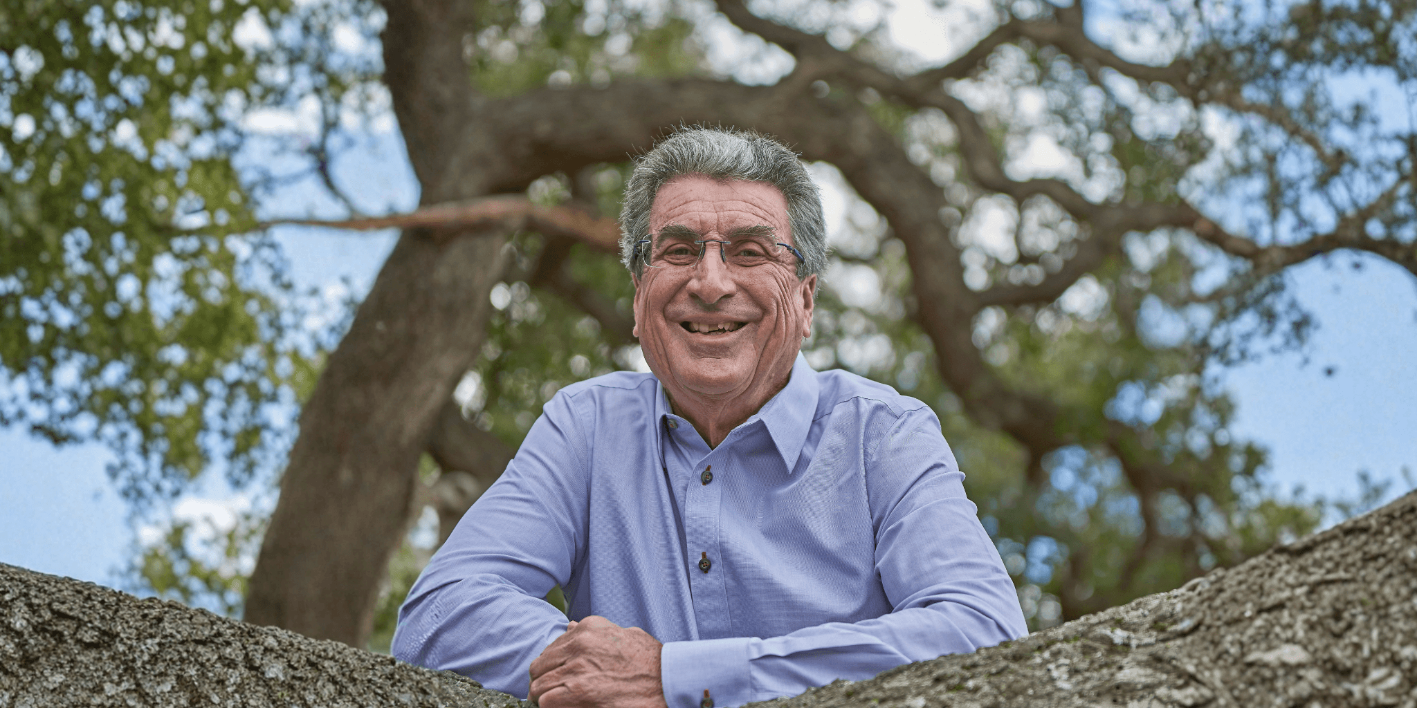 An older man in a light blue shirt smiles warmly, leaning on a tree branch. The background features a sunny, leafy setting with a blue sky, conveying a cheerful mood.