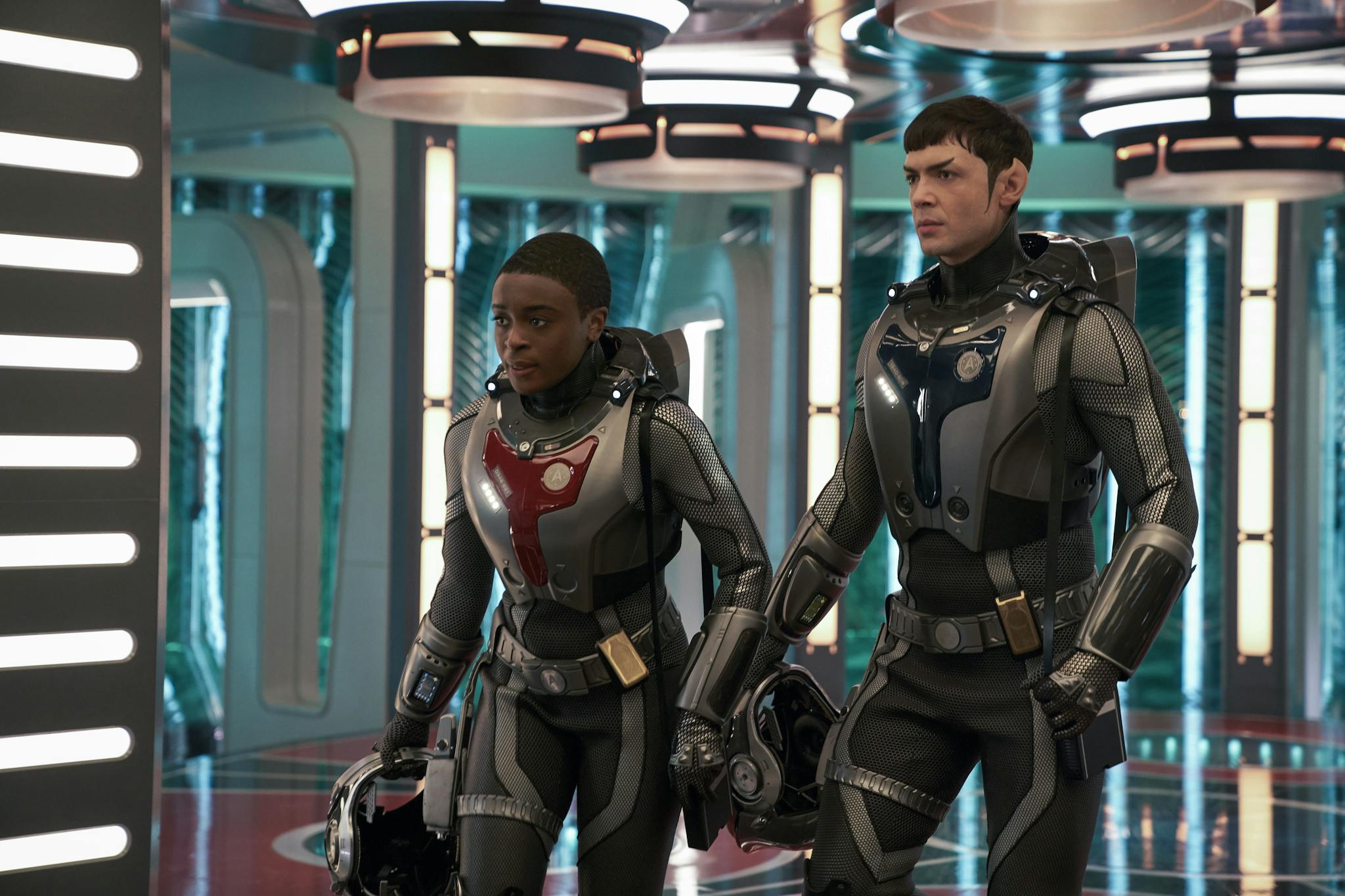 Two people in futuristic space suits walk through a high-tech corridor with circular lights on the ceiling. They carry helmets and appear focused on their mission. The setting is sleek and metallic, suggesting a sci-fi environment.