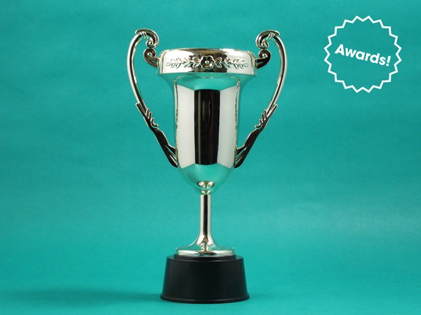 A shiny silver trophy with two handles and a black base stands against a teal background. A white starburst shape in the upper right corner contains the word "Awards!" in white text.