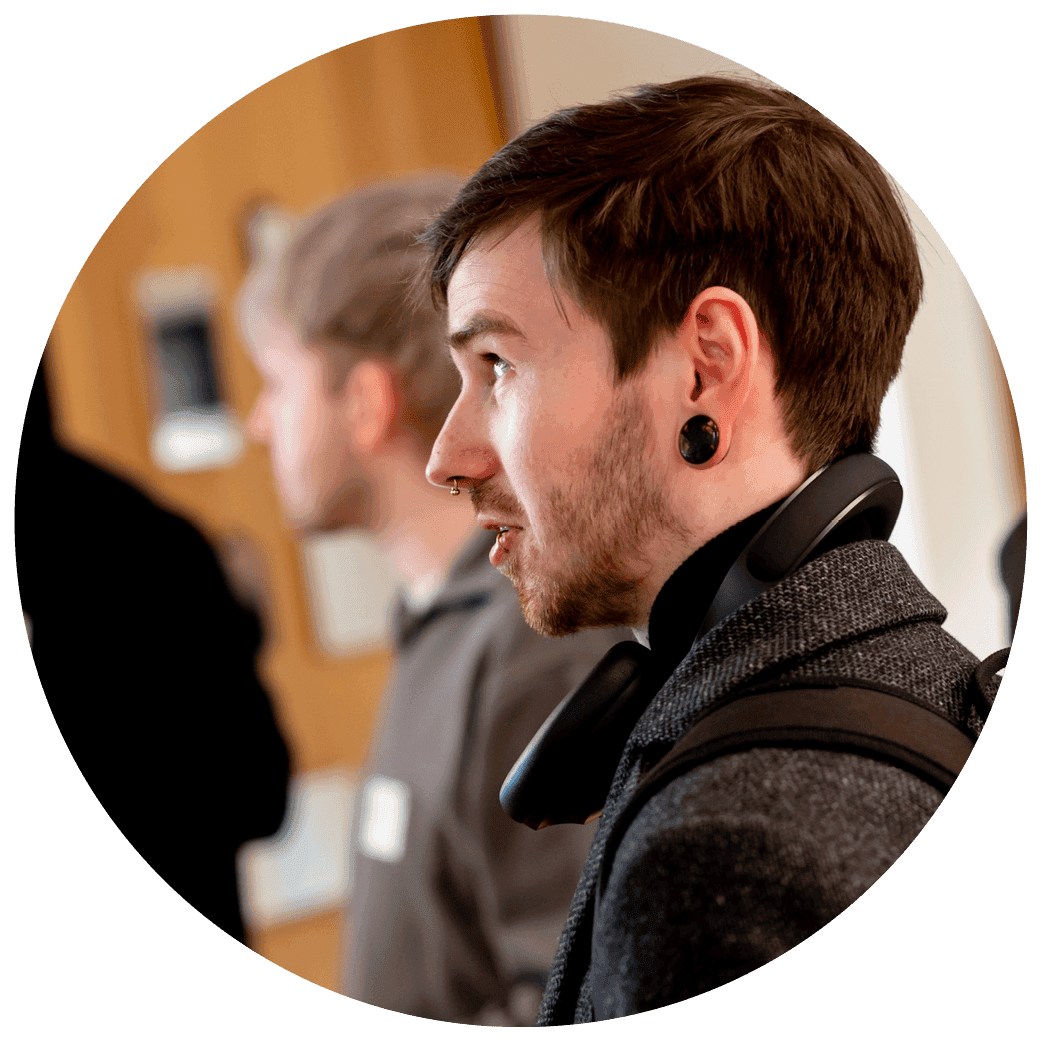 Person standing in a busy indoor setting wearing headphones around the neck and a dark coat, shown in profile.