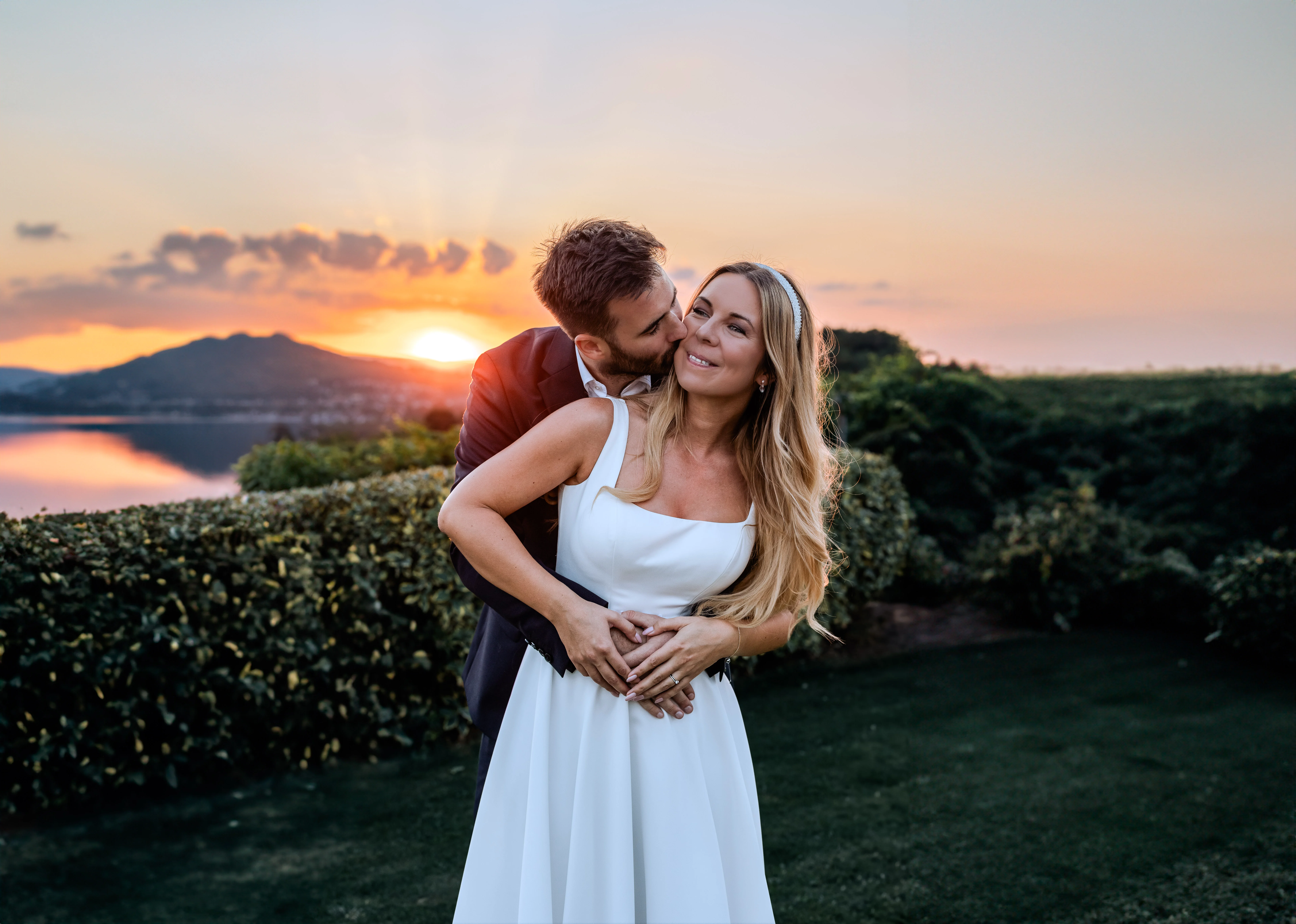 Esküvői ruhás pár romantikusan öleli egymást, háttérben Balaton naplementével, elegáns kertben.