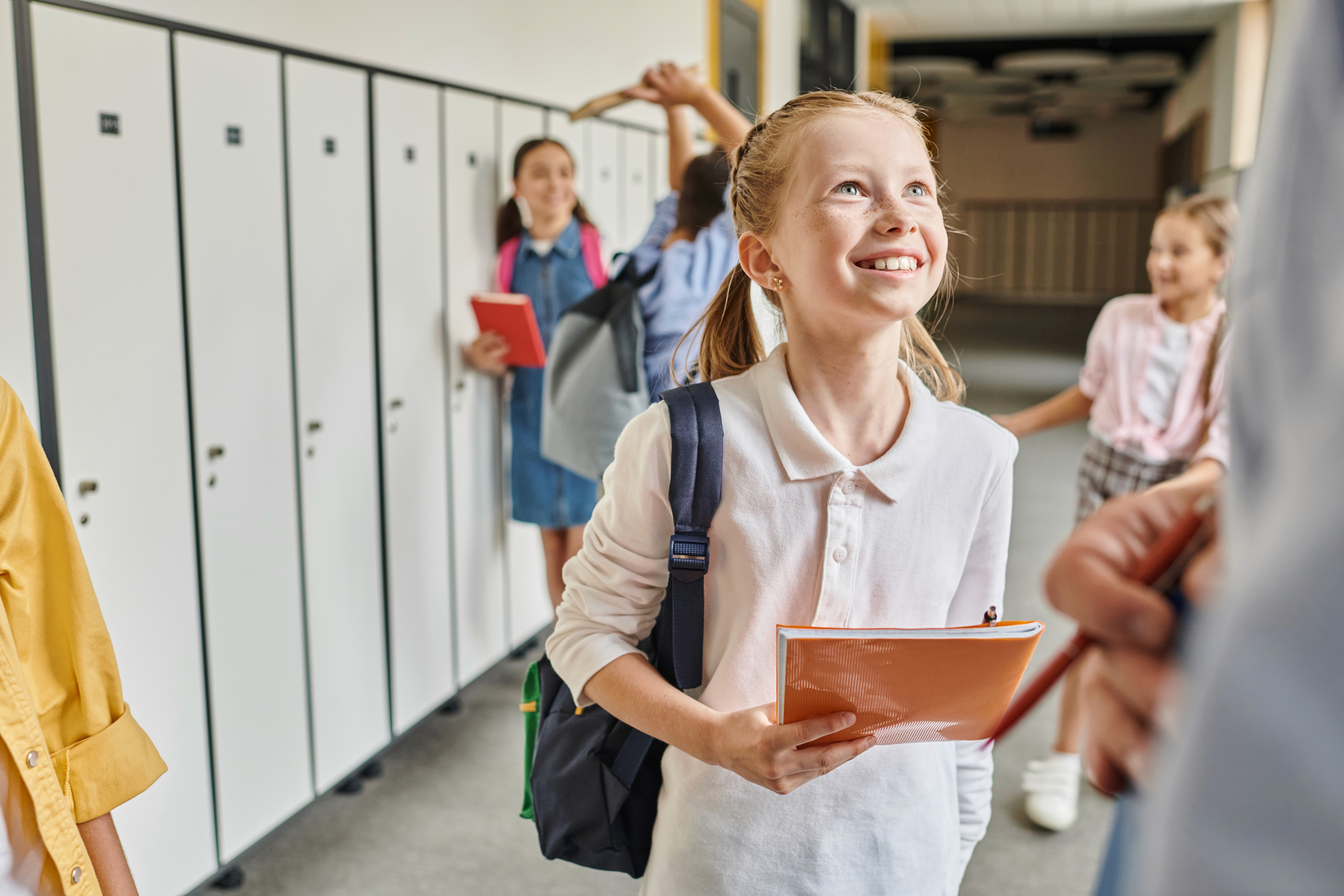 Fröhliches Schulkind mit Rucksack und Heft steht im Schulflur vor Schließfächern und startet motiviert in den Schultag