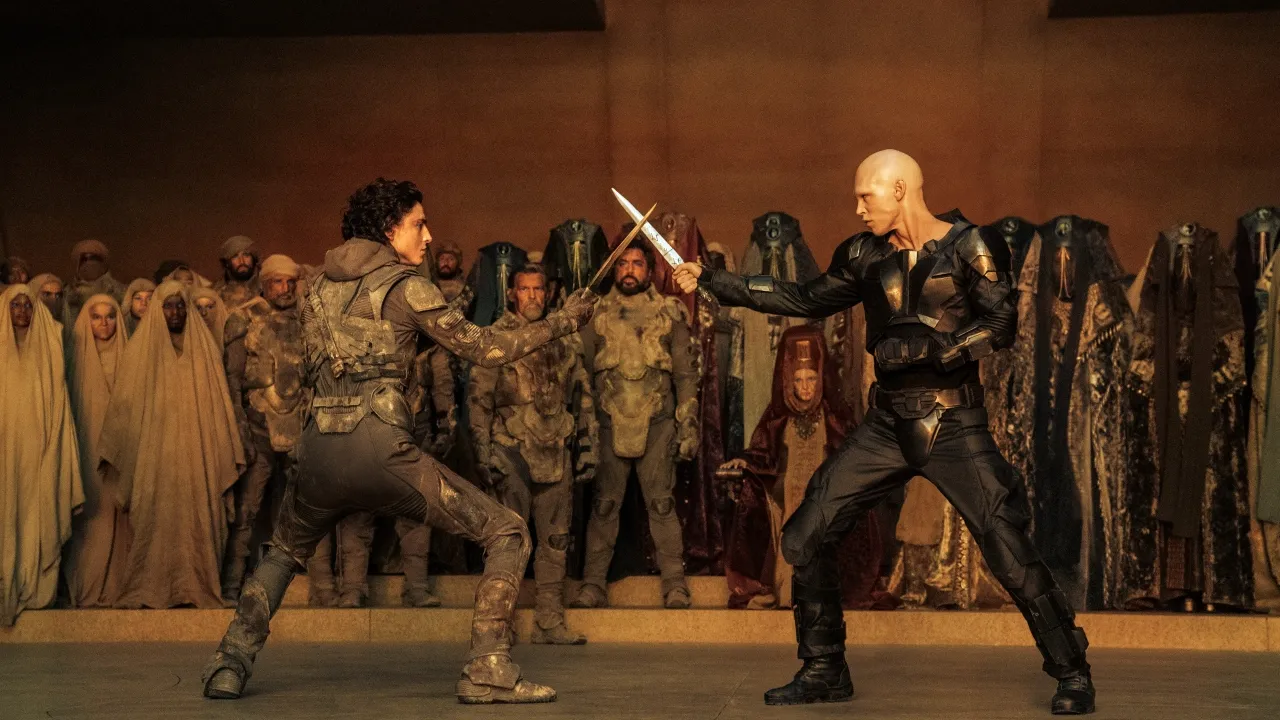 A dramatic scene depicts two figures in combat gear engaged in a tense knife fight, surrounded by an audience dressed in elaborate, otherworldly costumes, set against a grand, architectural backdrop.
