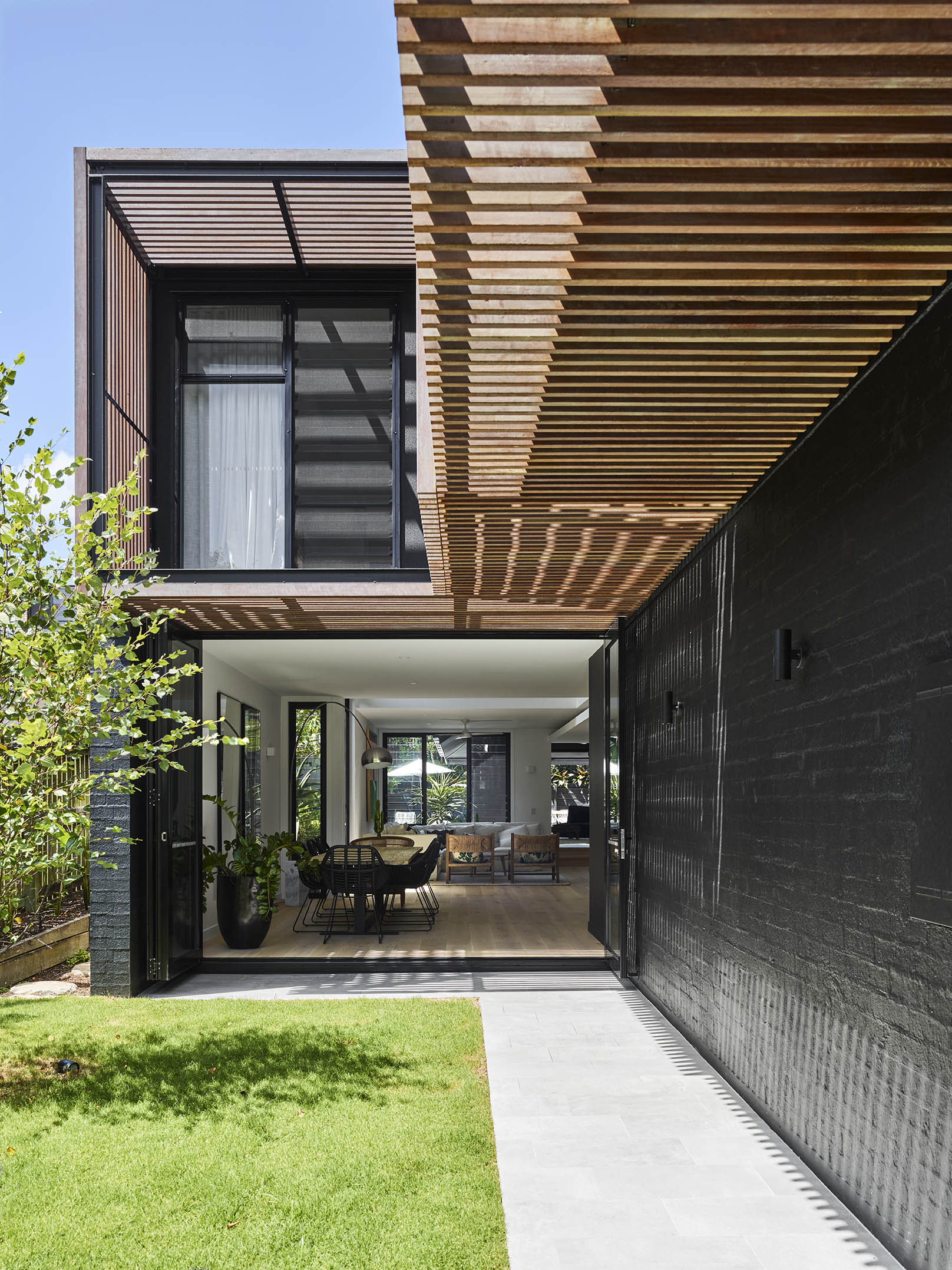 Open-plan indoor-outdoor living area with timber slatted ceiling, black brick wall, and garden outlook.