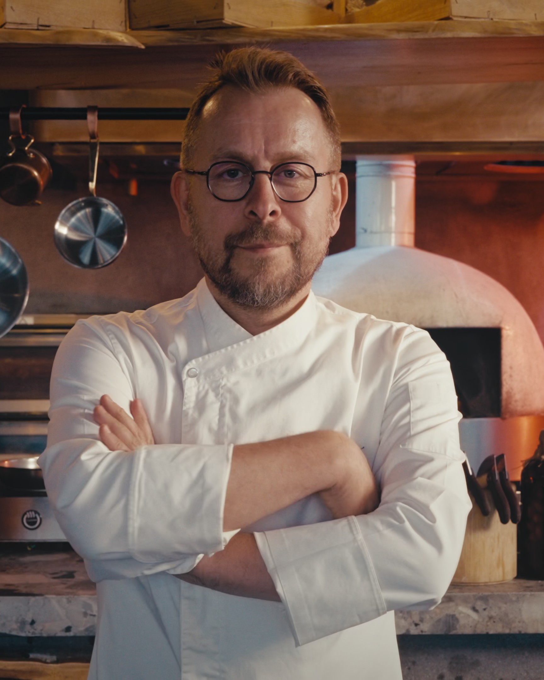 A chef in a white jacket standing with his arms crossed in his kitchen, copper pans and jars of preserves behind him