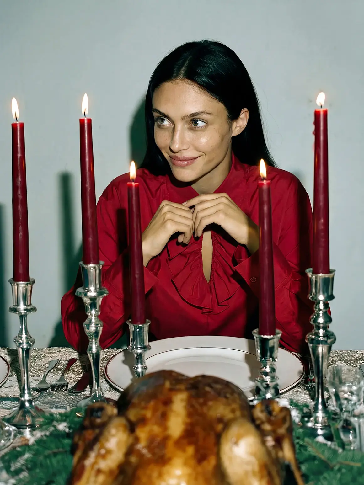 A woman in a red blouse sits at an elegantly set dining table with a roasted turkey centerpiece, surrounded by four tall red candles in silver holders, creating a high-end visual ambiance for a sophisticated fashion brand photoshoot.