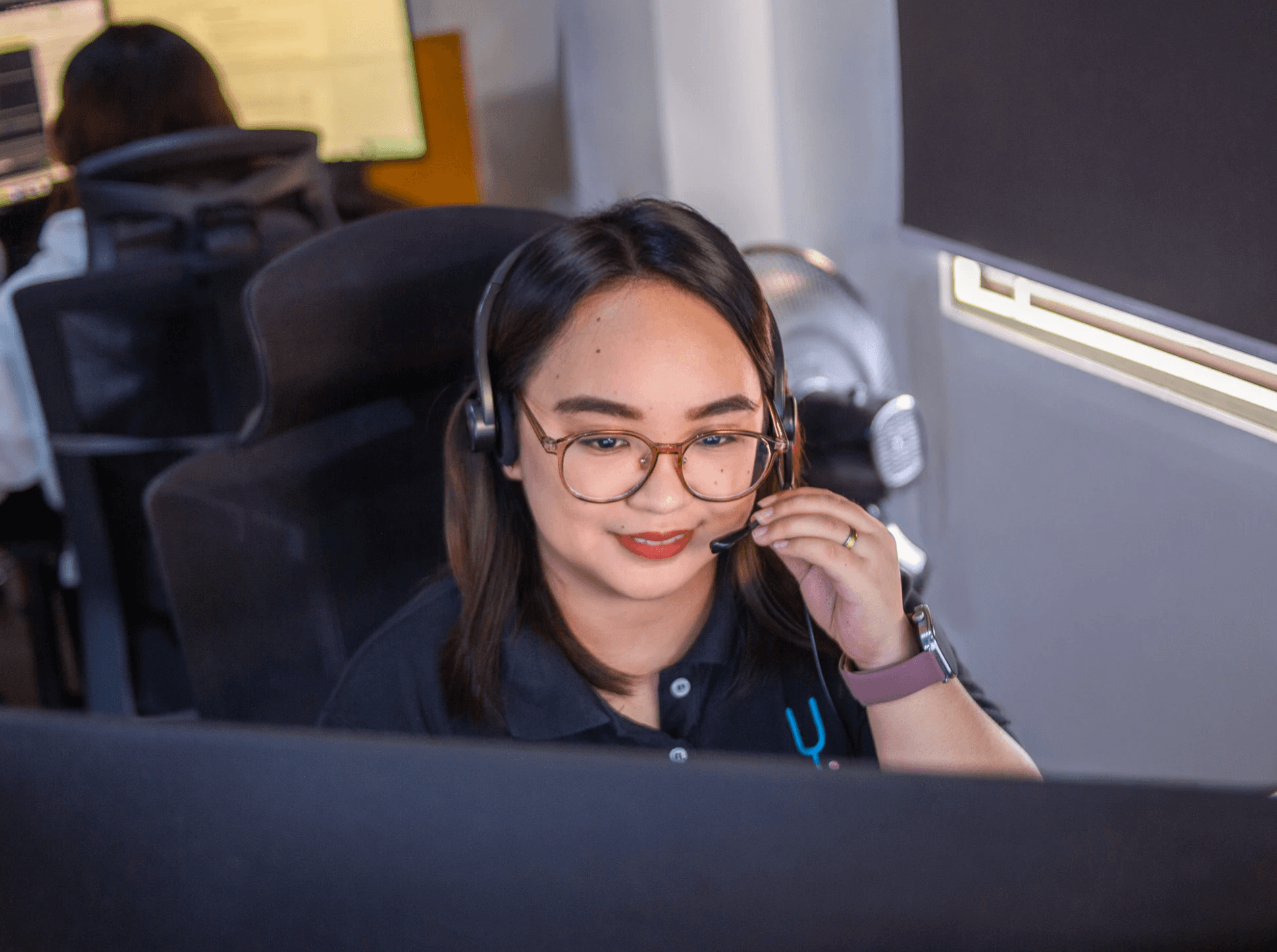 A woman wears glasses and smiles while working at a computer in a dimly lit workspace.