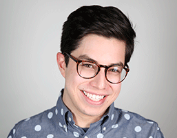 The image shows a young man with dark hair and glasses wearing a blue shirt with white polka dots. He is smiling and looking directly at the camera against a light gray background.