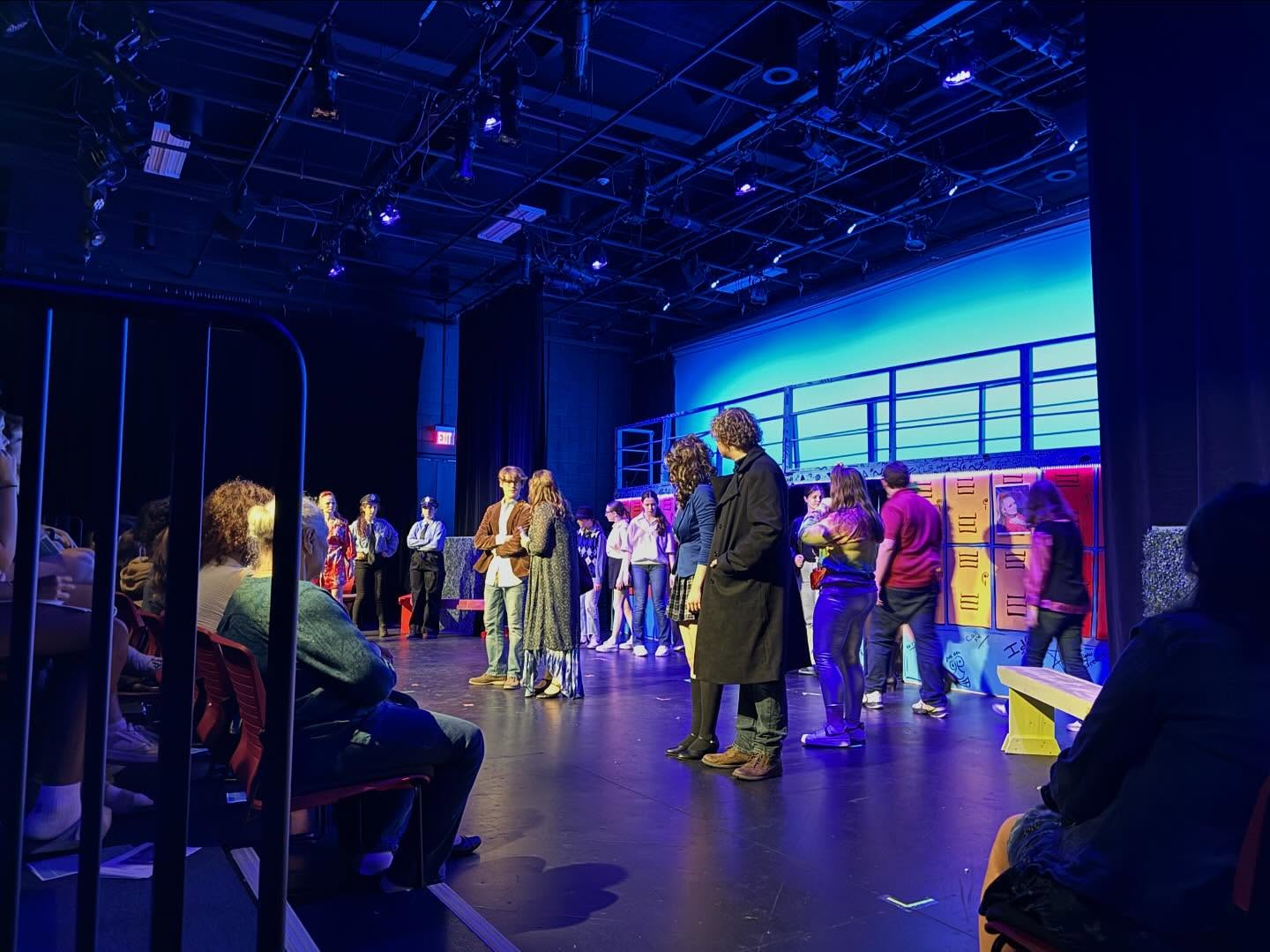 A scene from Heathers: Teen Edition in a blackbox theater. JD, Veronica, and the ensemble watch as Principal Gowan and Ms. Fleming speak under blue and purple lighting.