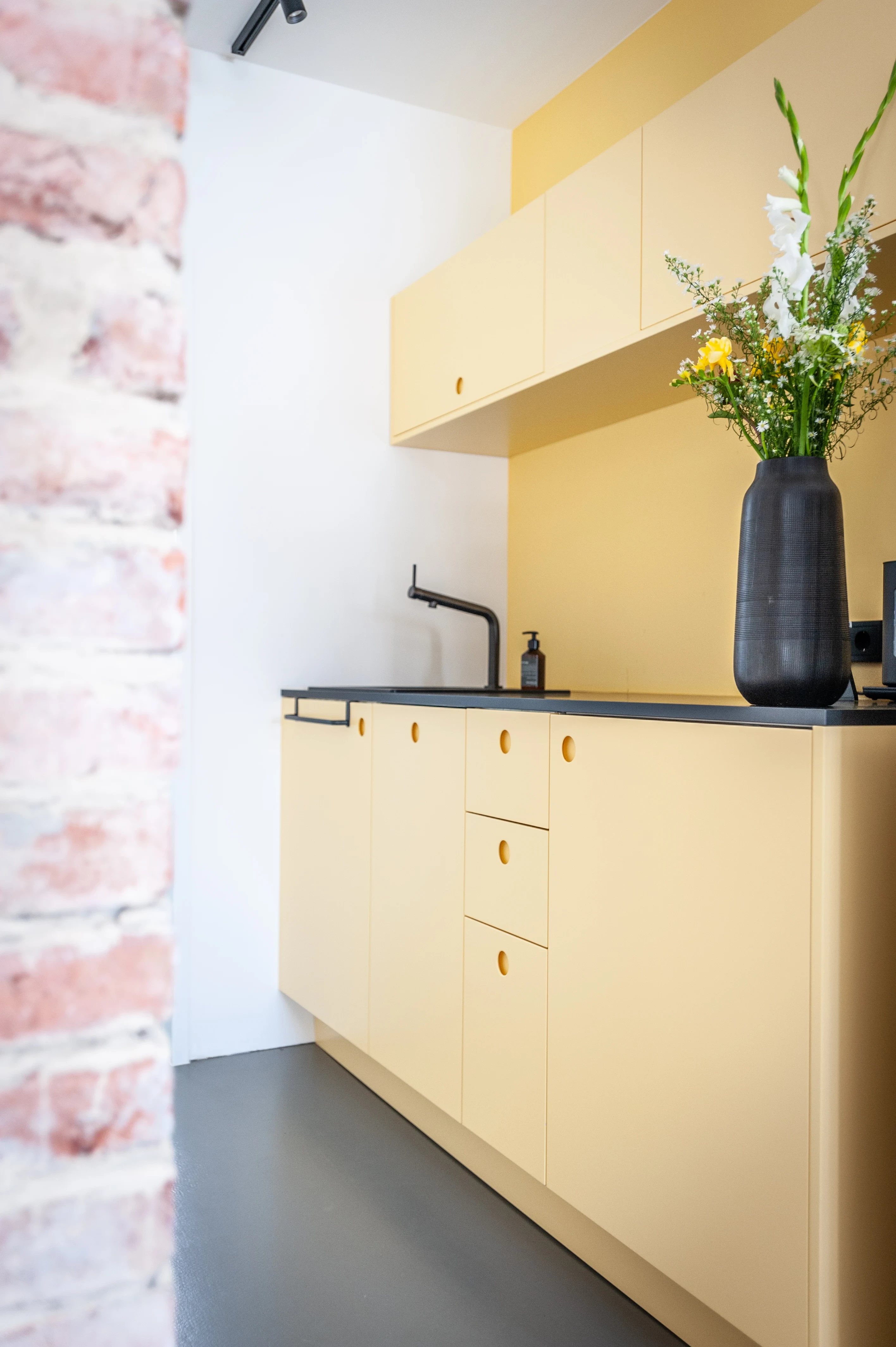 Small tea kitchen at EHEALTH-TEC with pale yellow cabinets with round cut-out handles, a black countertop and tap, a dark vase with fresh flowers and an exposed brick wall edge.