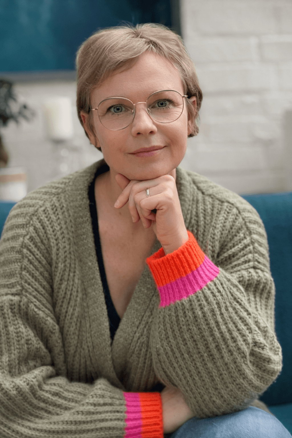 Smiling female therapist with short gray hair, wearing a brown top and a purple shawl, surrounded by a cozy background.