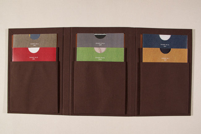 The image shows a triptych of dark brown leather-bound books mounted on a light beige wall. Each book has a rectangular front cover divided into three horizontal sections by horizontal stitching. The left section contains a red rectangle with a white circle in the center. The middle section contains a green rectangle with a white circle in the center. The right section contains a blue rectangle with a white circle in the center. Each cover also has a small, circular cutout in the top center. The books are arranged in a row, with each cover slightly offset from the one to its left.