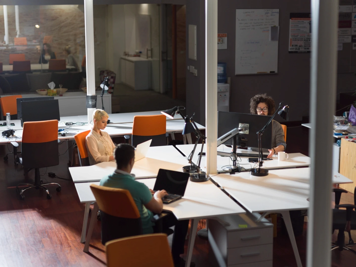 A modern, open-plan office space with several people working at white desks equipped with computers and office lamps, featuring stylish orange chairs and a warm wooden floor.
